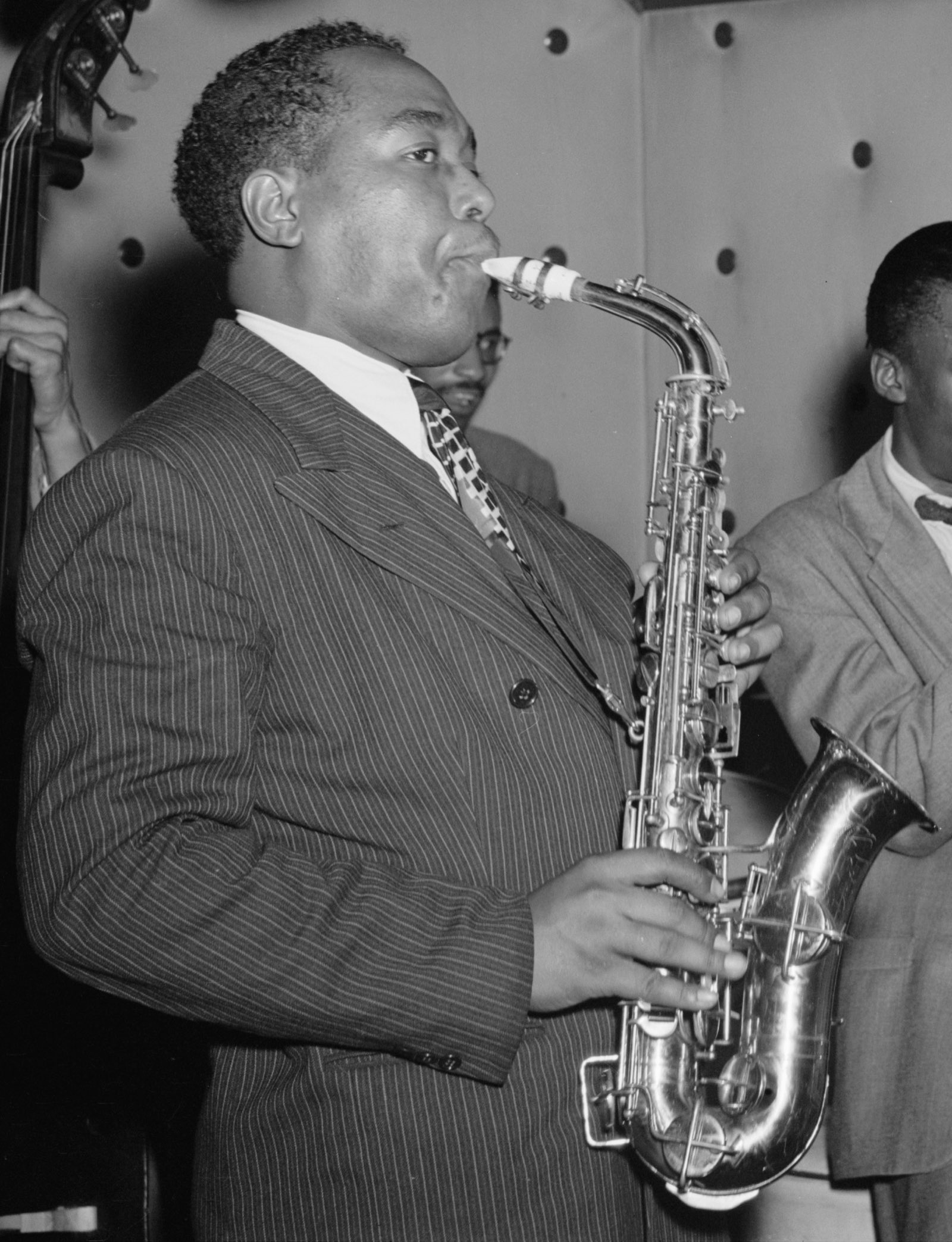 Charlie Parker de perfil tocando saxofone alto, de terno risca de giz, com um contrabaixo ao fundo.