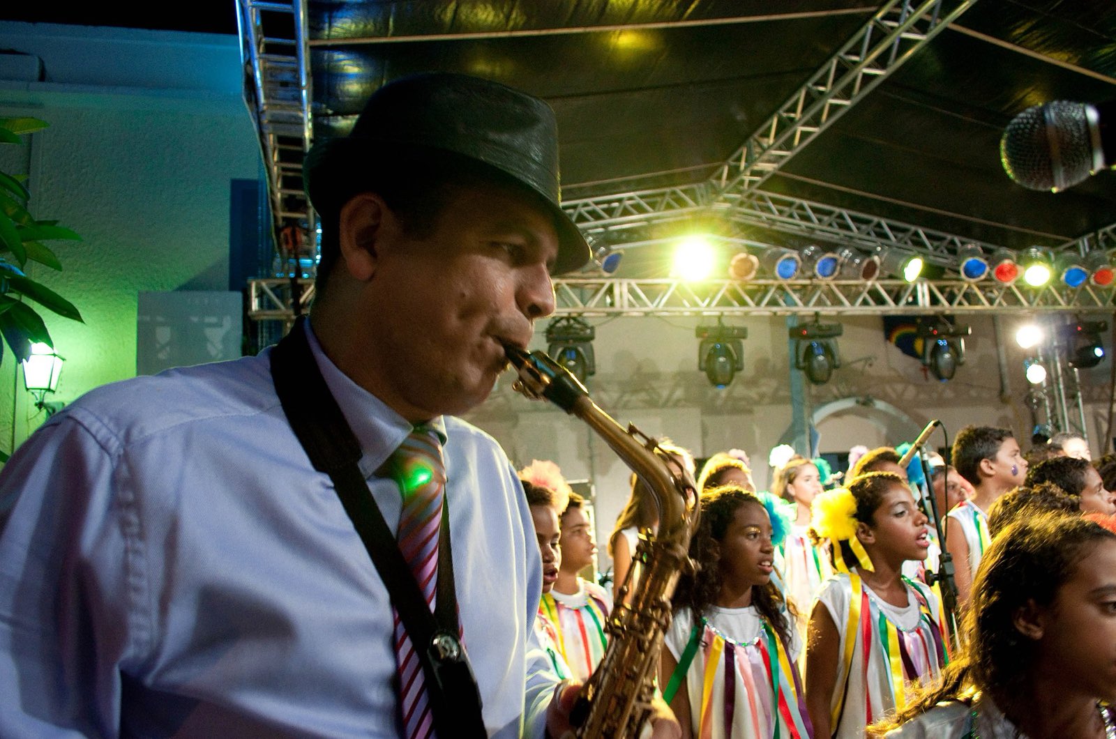 Maestro Spok tocando saxofone num palco de rua em Olinda, com crianças fantasiadas ao fundo.