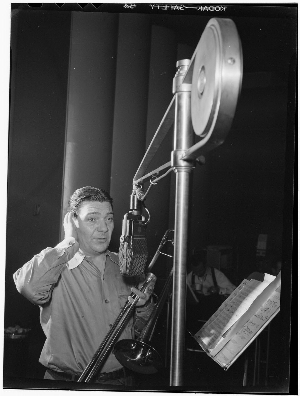 Jack Teagarden no estúdio, com a mão no ouvido e o trombone apoiado no braço, diante de um microfone.
